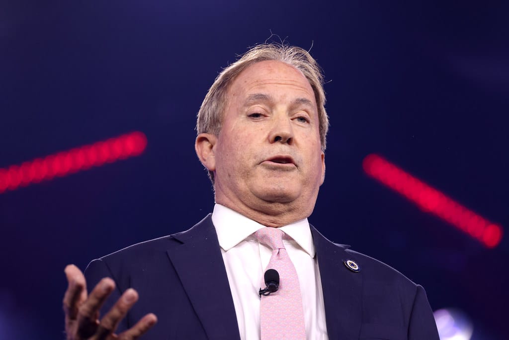 Texas Attorney General Ken Paxton speaks on stage in a suit and pink tie, with red lights in the blurred background.